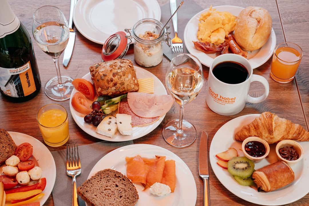 Ein Frühstücksbuffet auf einem Holztisch mit verschiedenen Brotsorten, Räucherlachs, Käse, Obst, Joghurt, Kaffee, Orangensaft und Wein. Ein Croissant mit Marmelade und ein Brötchen mit Speck runden das Angebot ab.
