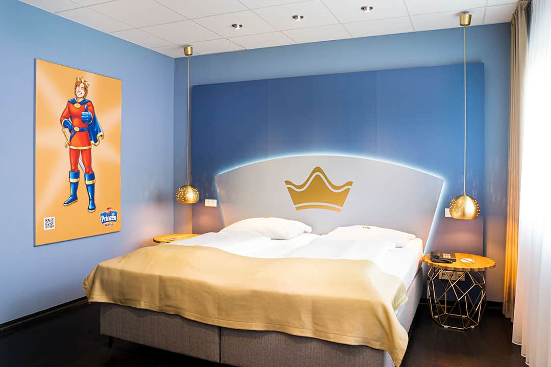 Ein Hotelzimmer mit einem Doppelbett mit goldenem Kopfteil, Nachttischen mit Hängelampen und blauen Wänden. An der Wand hängt ein großes Poster einer Zeichentrickfigur in rot-blauem Outfit. Tageslicht fällt durch transparente Vorhänge.