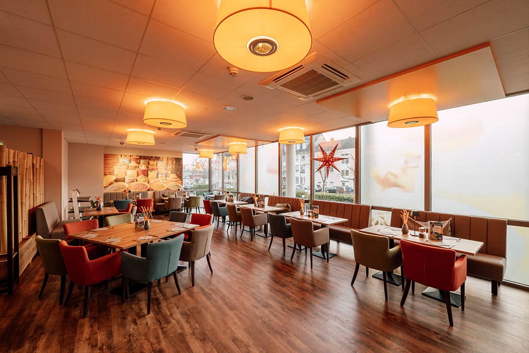 Ein geräumiges, warm beleuchtetes Restaurant mit Holzboden und modernen Pendelleuchten. Die Tische sind von bunten Polsterstühlen umgeben. Große Fenster säumen eine Wand und lassen natürliches Licht herein.