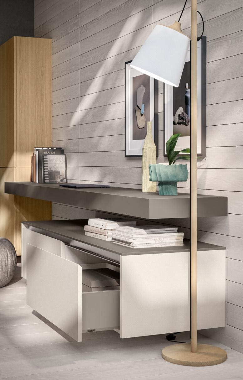 A modern interior with a wooden floor and wall, featuring a floating shelf with decorative items and books. Below is a drawer unit with open drawers. A floor lamp in the foreground casts light on the scene.