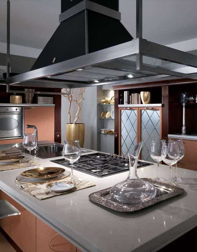 Modern kitchen with a central island featuring a gas stove, sink, wine glasses, and a decanter on a silver tray. The space has sleek cabinets, a metallic range hood, and decorative elements.