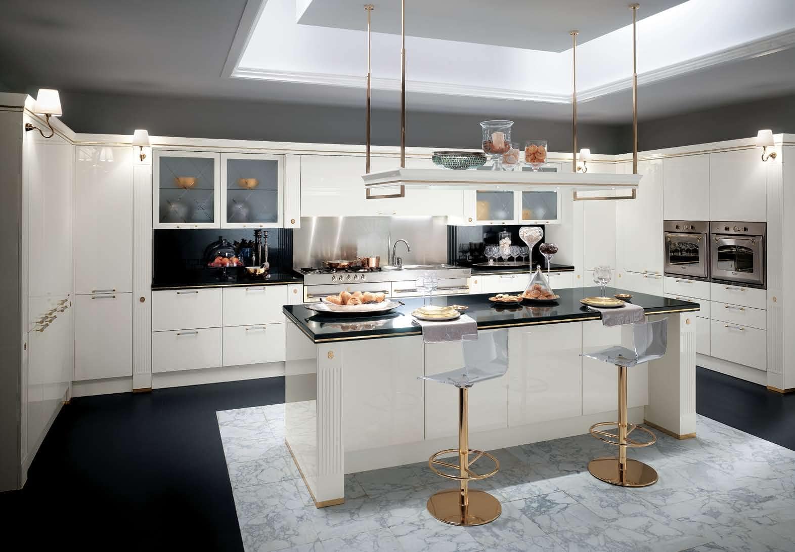 Modern kitchen with white cabinets, black countertops, a central island, two transparent bar stools with gold bases, stainless steel appliances, and overhead lighting.