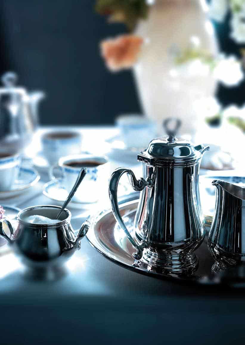 Silver tea set with teapot, sugar bowl, and creamer on a tray, surrounded by cups and saucers on a table.