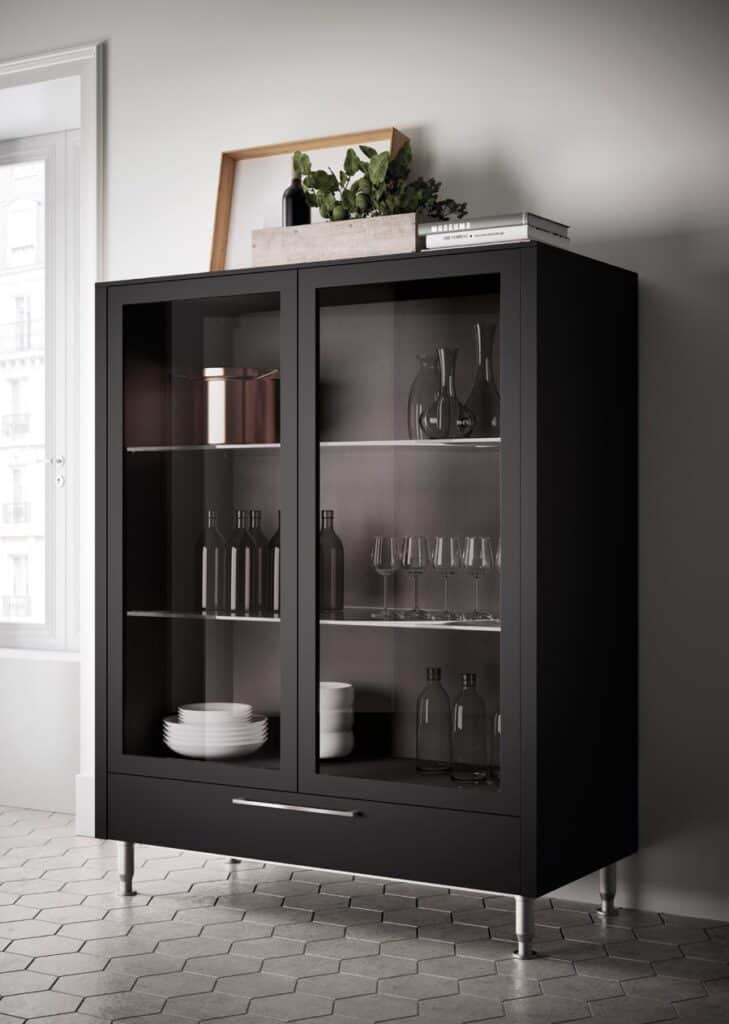Black cabinet with glass doors showcasing glassware, bottles, and stacked plates, set in a bright room with hexagonal floor tiles. A plant and books decorate the top of the cabinet.