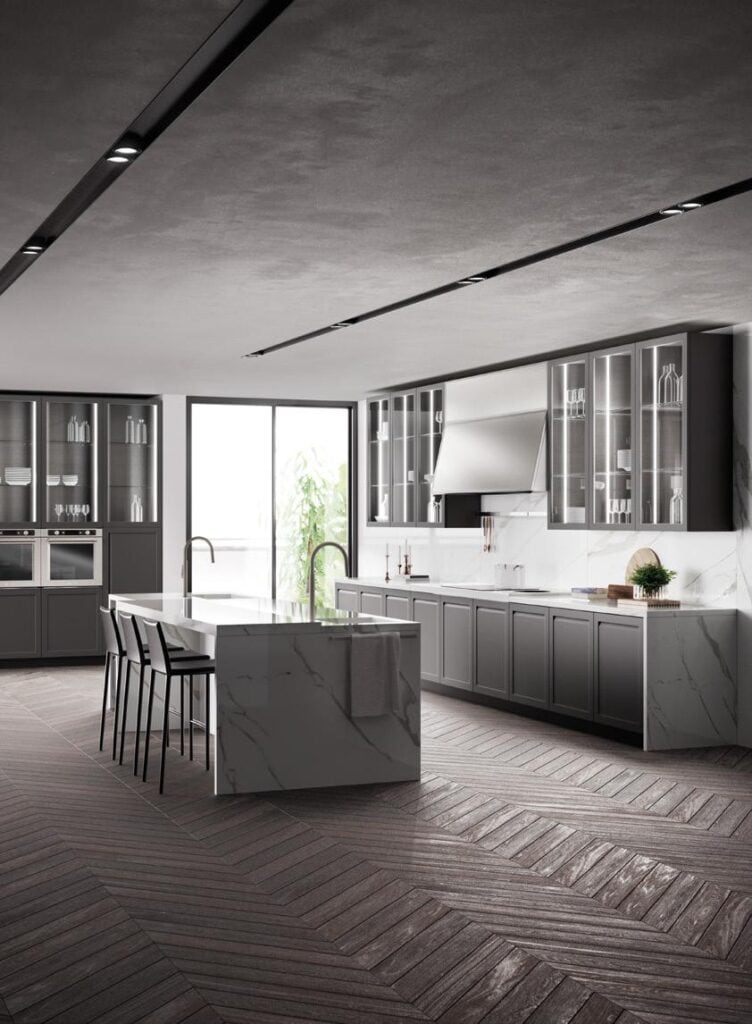 Modern kitchen with gray cabinetry, marble countertops, a central island with bar stools, and herringbone-patterned wood flooring.