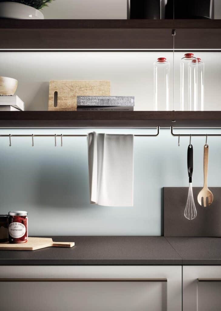 Modern kitchen shelves with glass jars, a cutting board, and books; below are hanging utensils and a towel rail.