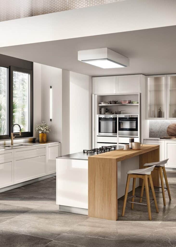 Modern kitchen interior with white cabinets, large window, central island with wooden counter, stools, built-in ovens, and cooktop.