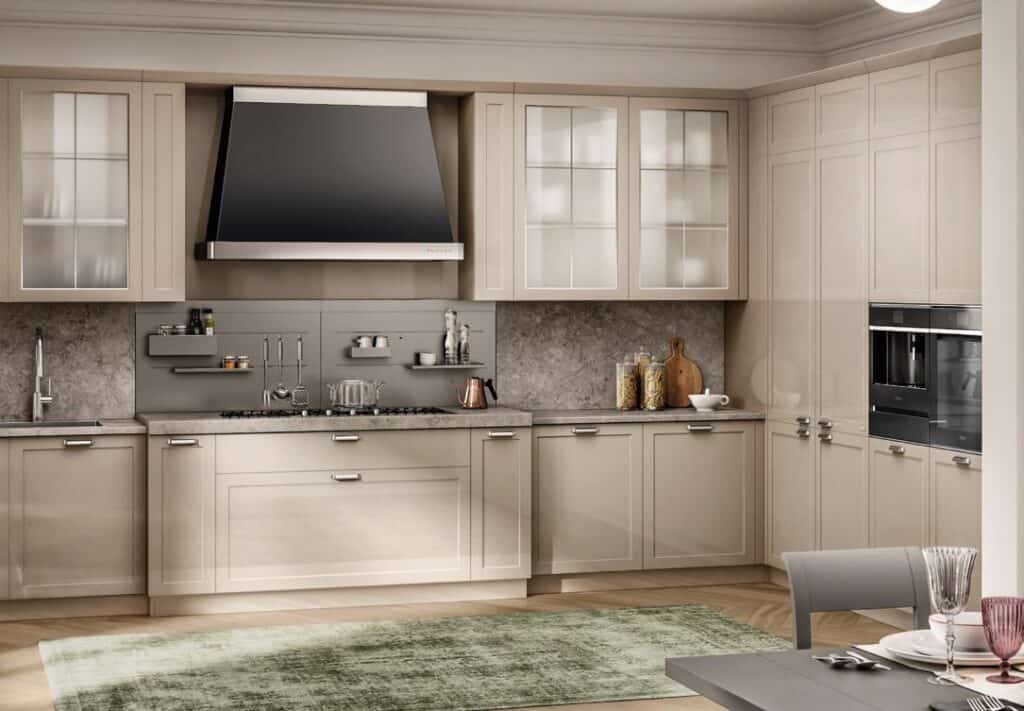 Modern kitchen with light beige cabinets, glass-front upper cabinets, a stainless steel range hood, integrated oven, and marble countertops. A green rug and dining table are visible.