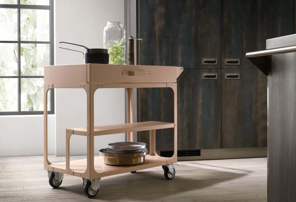 A beige kitchen cart with wheels, featuring a jar of herbs and kitchen utensils on top, is placed in a modern kitchen with dark cabinets and a large window.