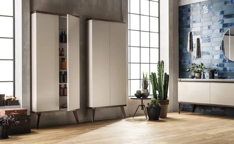 Modern living room with two tall cabinets, one partially open revealing bottles, wooden floor, potted plants, blue tiled wall, round mirror, and decorative items on a sideboard.