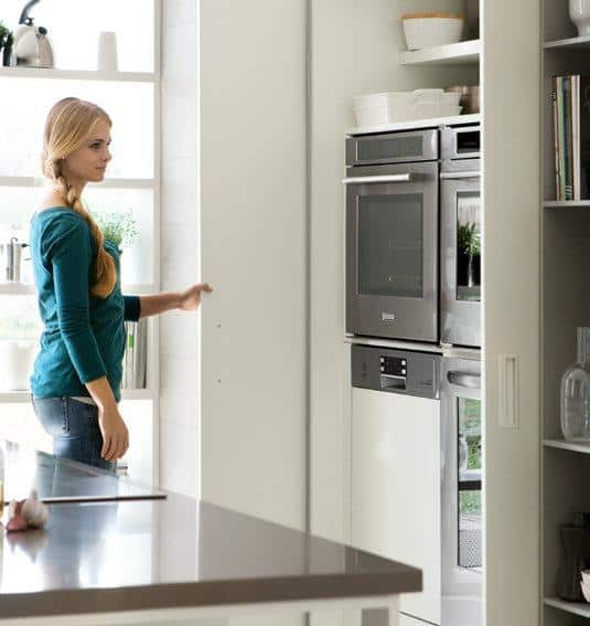 Woman standing in a kitchen with modern appliances and open shelving.