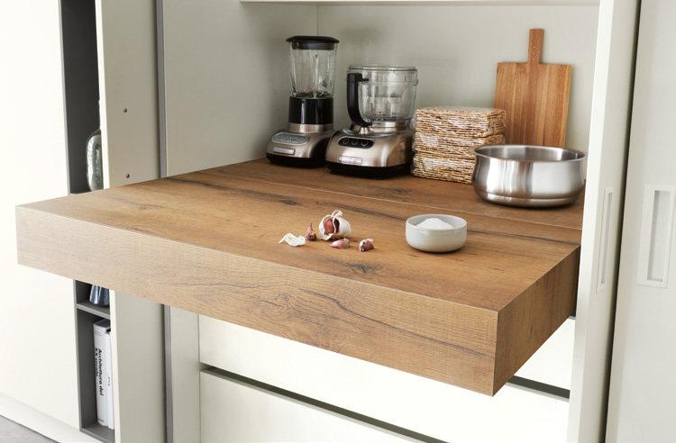 Wooden kitchen countertop with a blender, food processor, cutting board, stacked kitchen towels, stainless steel bowl, garlic cloves, and a small bowl.