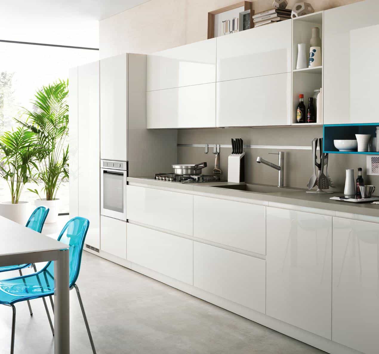 Modern kitchen with glossy white cabinets, stainless steel appliances, and a countertop. Blue transparent chairs at a dining table, large potted plant near a window.