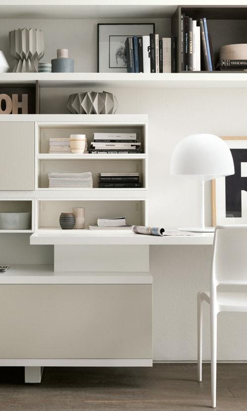 Modern home office setup with a sleek white desk, bookshelves, and a contemporary white chair. Decor includes books, a white lamp, and geometric vases.