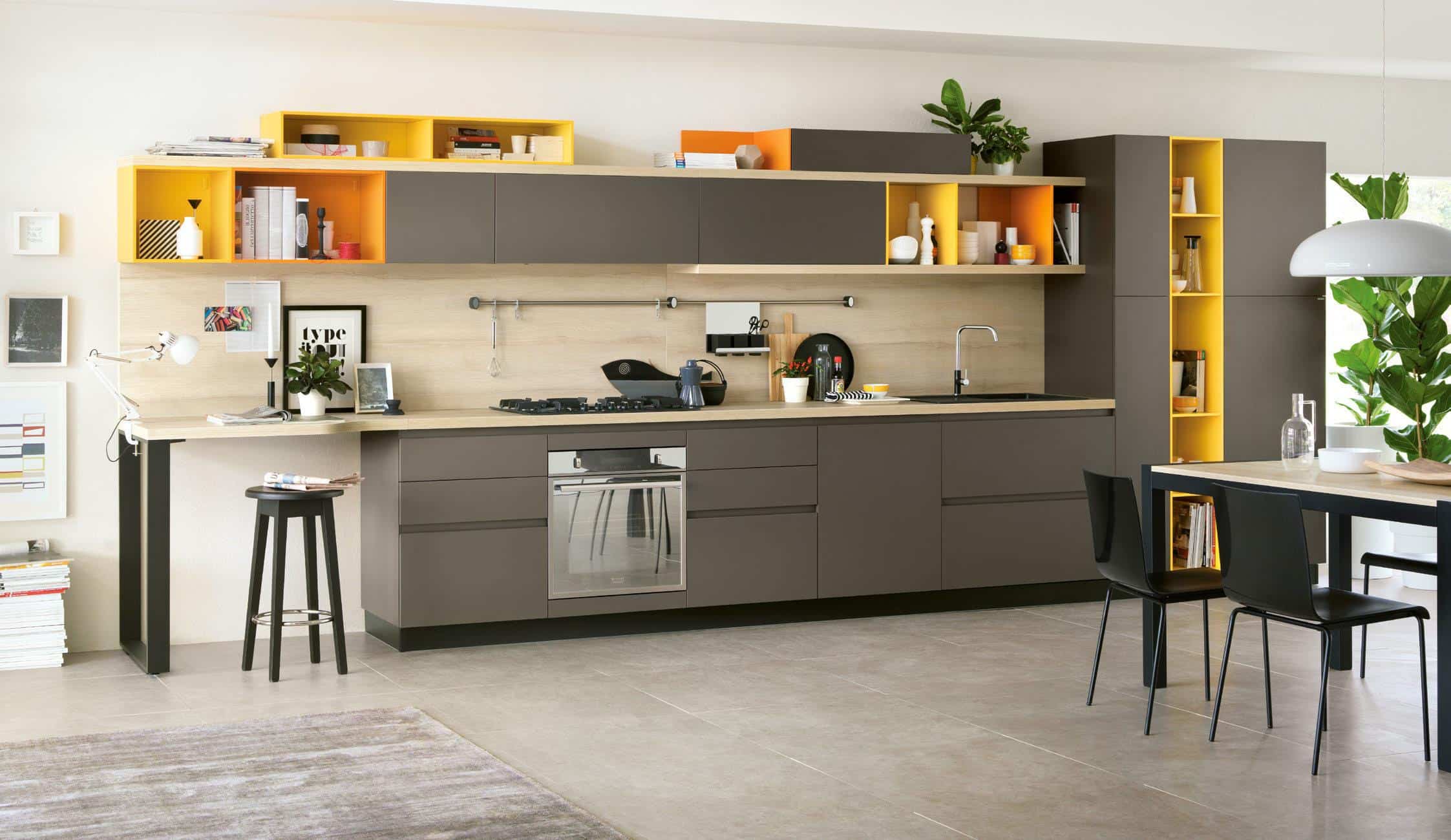 Modern kitchen with dark cabinets, yellow shelving accents, built-in oven, and a light wood countertop. A small black stool sits near a desk area, and a dining table with black chairs is on the right. Indoor plants add a touch of greenery.
