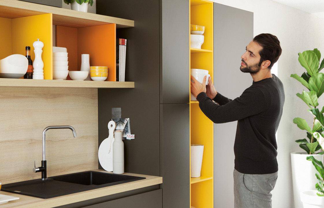 Man organizing dishes in a modern kitchen with open shelves and yellow accents.