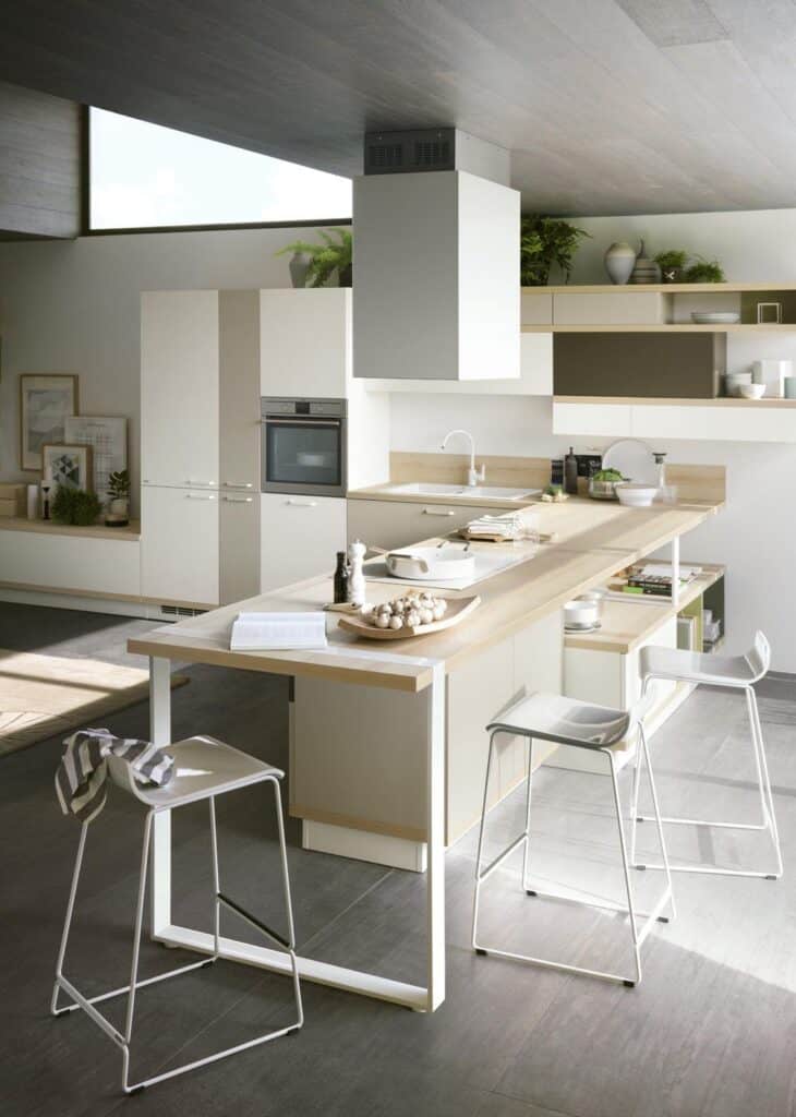 Modern kitchen with a white island, bar stools, and built-in oven, featuring light wood accents and greenery.