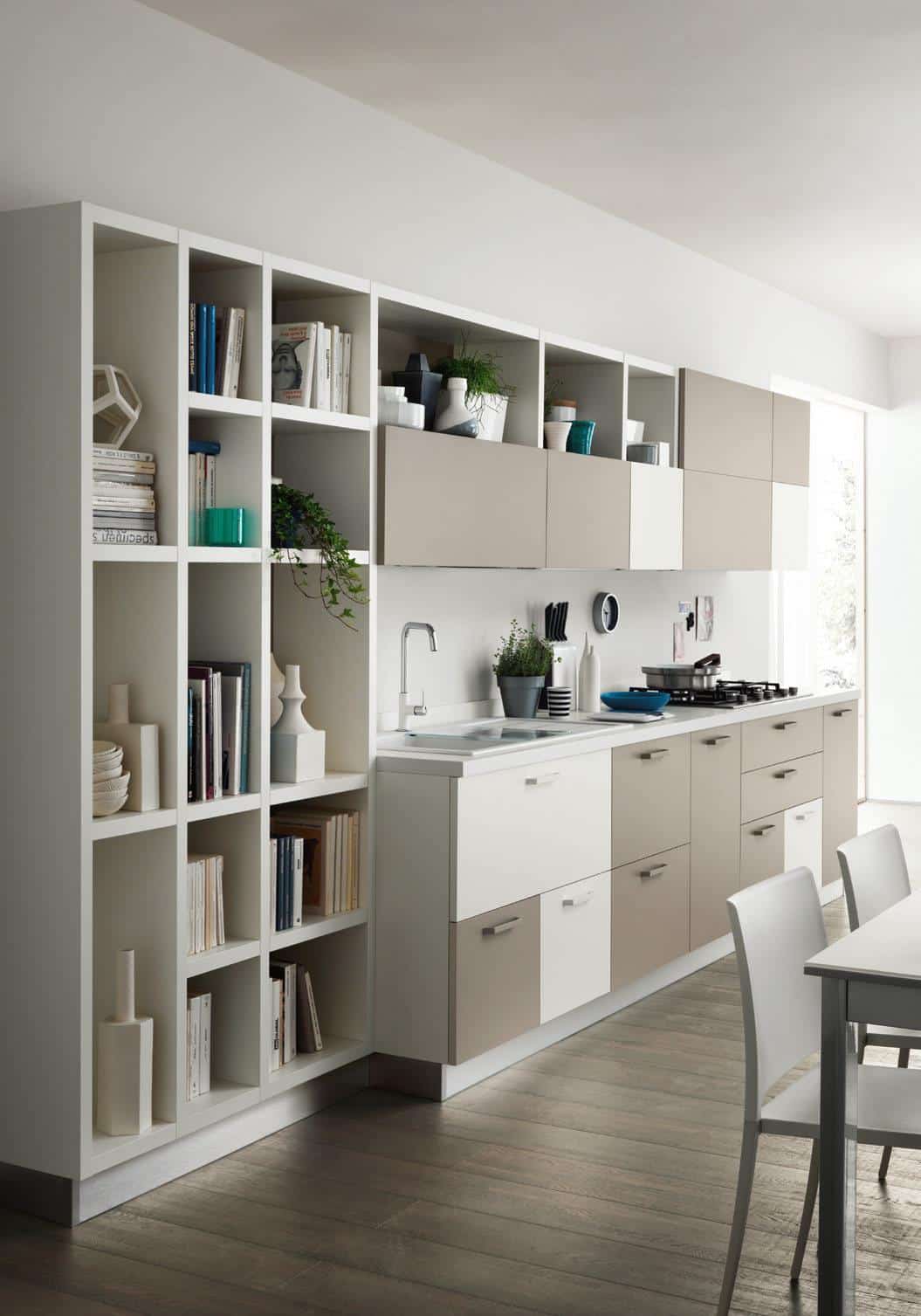 Modern kitchen with beige and white cabinets, a built-in shelving unit displaying books and decor, a sleek countertop with a sink and stove, and a dining table with chairs.