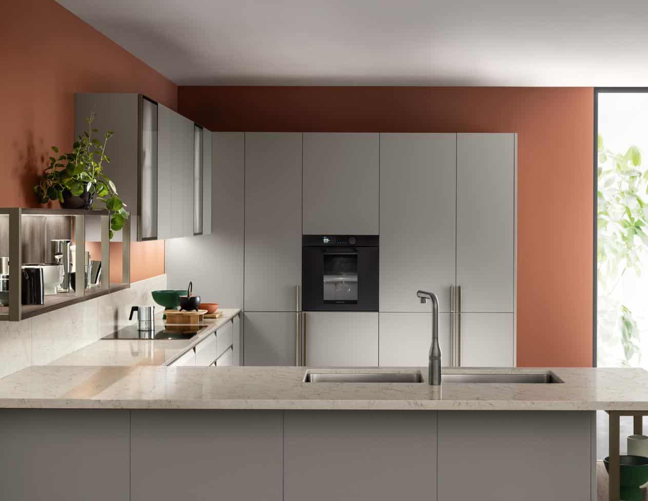 Modern kitchen with gray cabinets, a marble countertop, orange walls, and a built-in oven. A potted plant and kitchenware are on display, and there's a window showing greenery outside.
