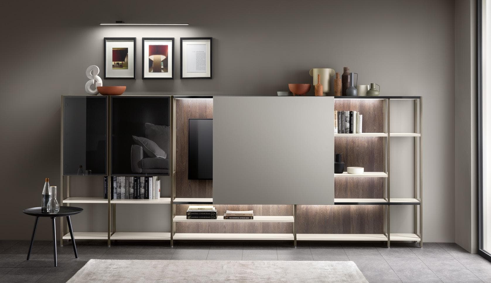 Modern living room with a sleek wall unit featuring open shelves, decorative items, books, and a mounted TV partly covered by a sliding panel, with a small round table displaying a bottle and glass in front.