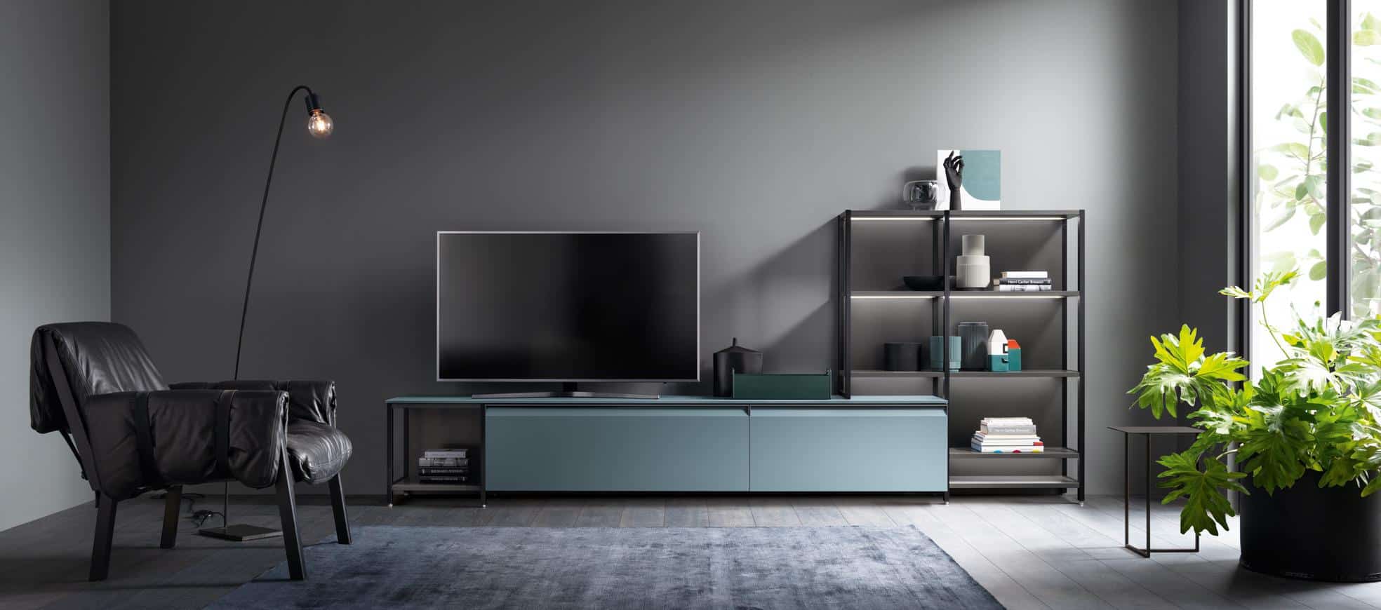 Modern living room with a black leather chair, flat-screen TV on a teal console, floor lamp, bookshelf with decor items, large potted plant, and gray walls.