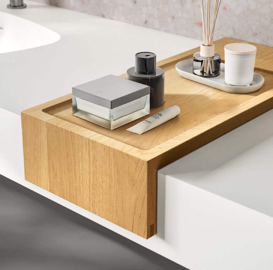 Wooden bathroom tray with toiletries including a square glass jar, a black bottle, a small tube, a diffuser, and a candle on a white countertop.