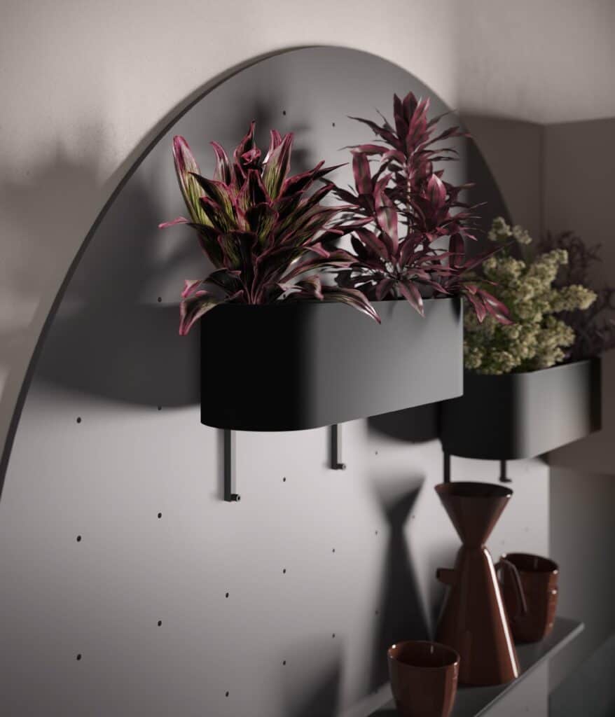 Indoor wall planter with vibrant pink and green plants, mounted on a curved, perforated panel; below are dark-colored ceramic vases and cups.