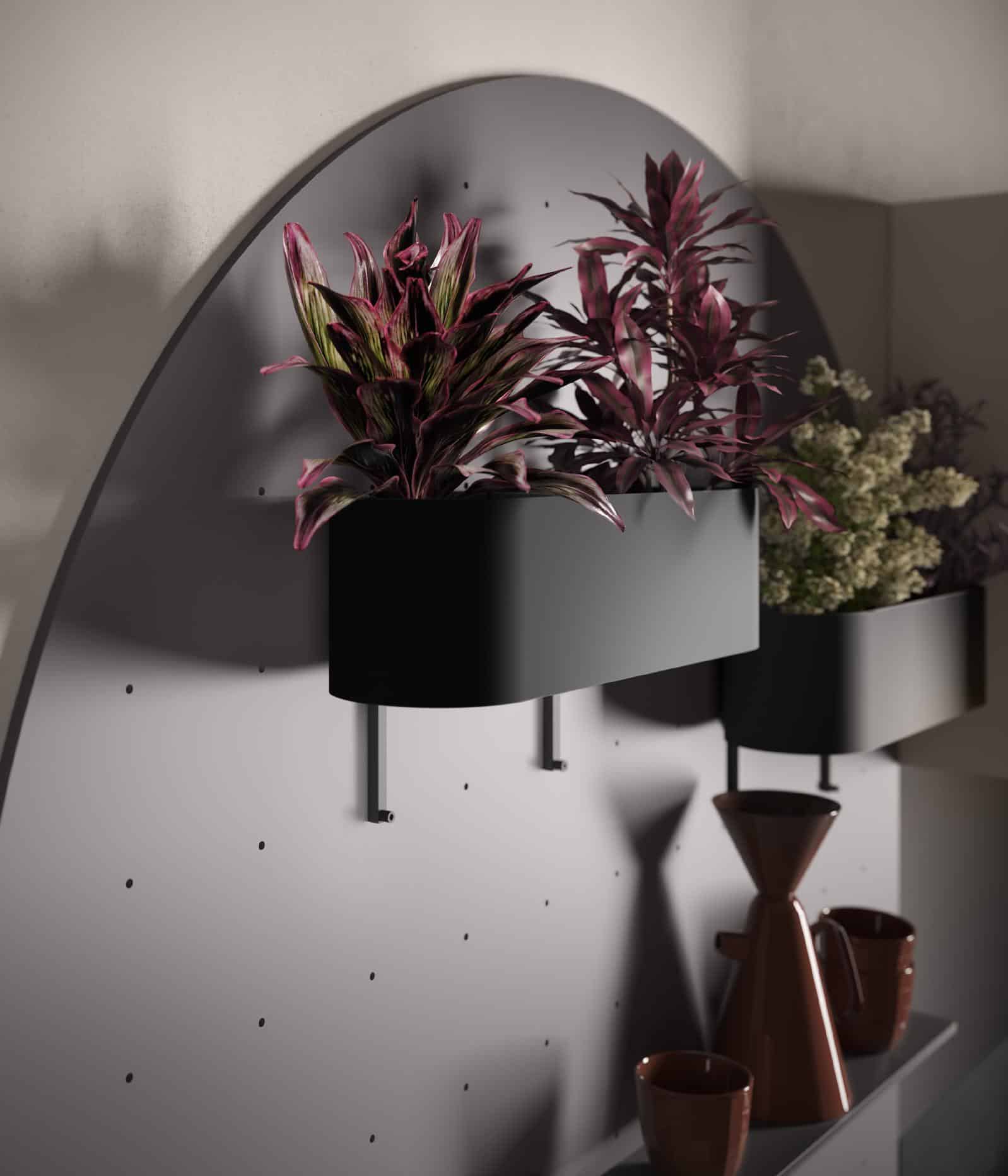 Indoor wall planter with vibrant pink and green plants, mounted on a curved, perforated panel; below are dark-colored ceramic vases and cups.