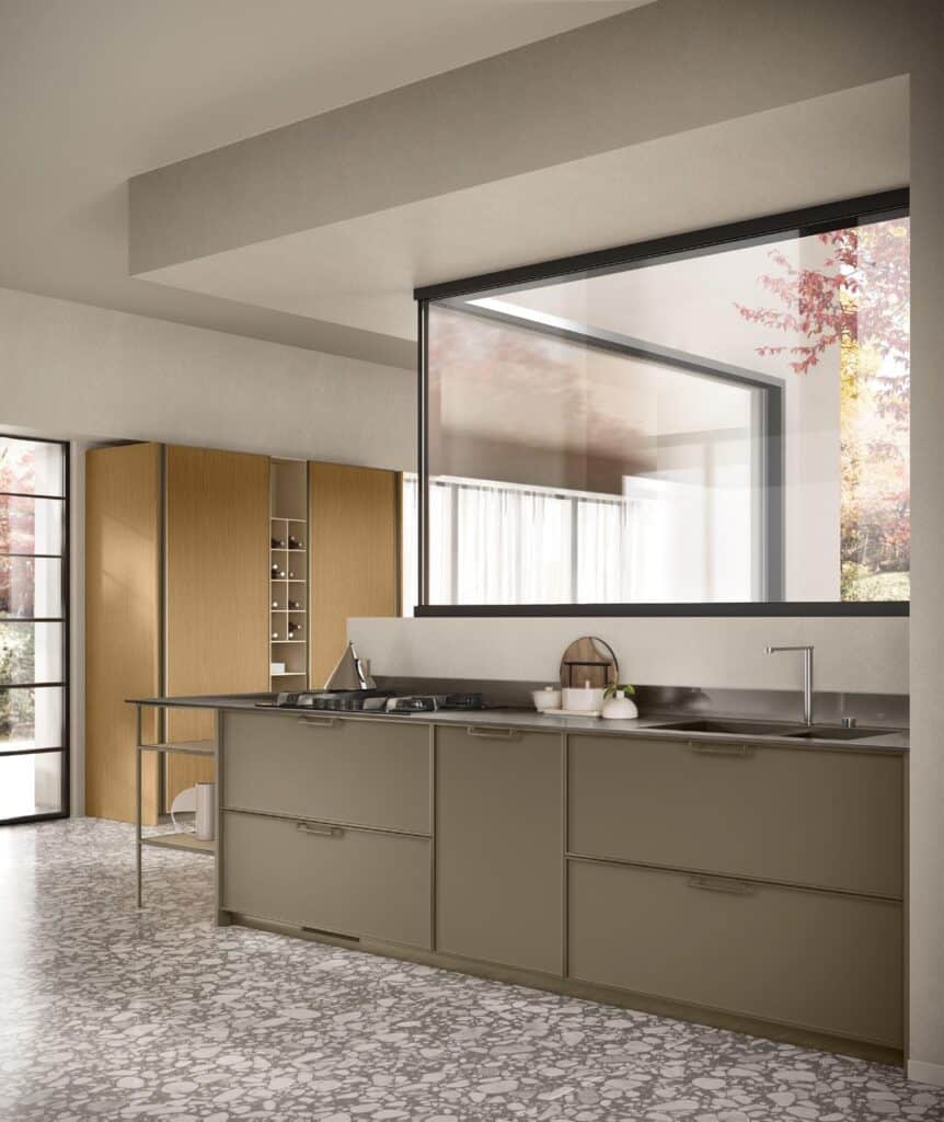 Modern kitchen with olive green cabinets, stainless steel countertop, and built-in stove. Large window with outdoor view and wooden cabinets on left. Light-colored terrazzo flooring.
