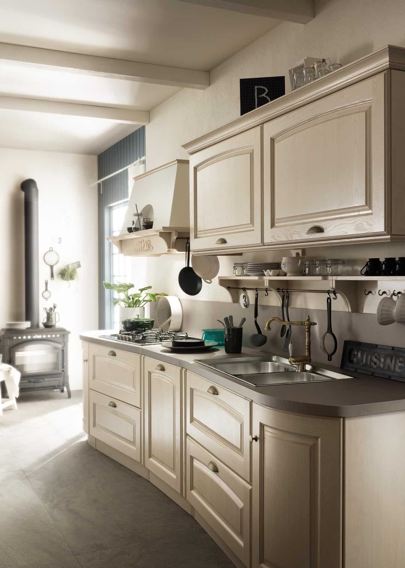 Traditional kitchen with light wood cabinetry, wall-mounted shelves, and utensils hanging above a sink. A stove is next to a decorative exhaust hood, with a small wood-burning stove in the corner.