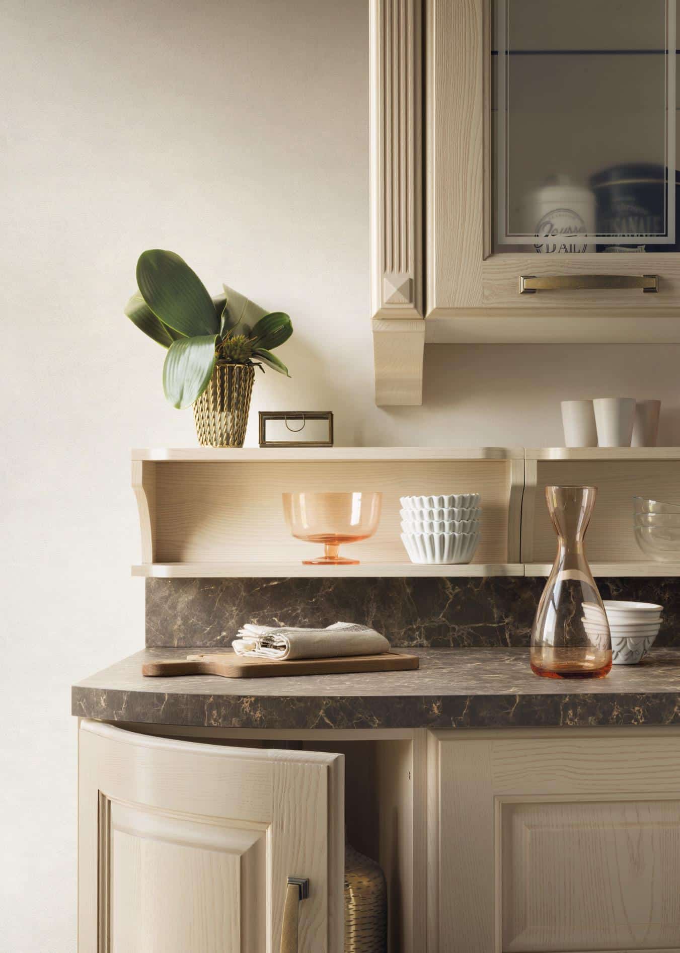 A kitchen countertop with a marble surface, featuring a wooden cutting board with folded cloths, a clear carafe, a decorative plant in a pot, and assorted dishware on shelves.