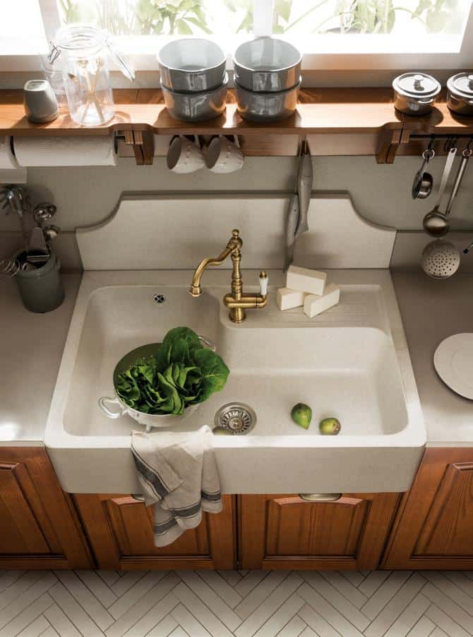 Kitchen sink with brass faucet, lettuce in a colander, two avocados, three soap bars, and a towel. Above are mugs and bowls on a wooden shelf.