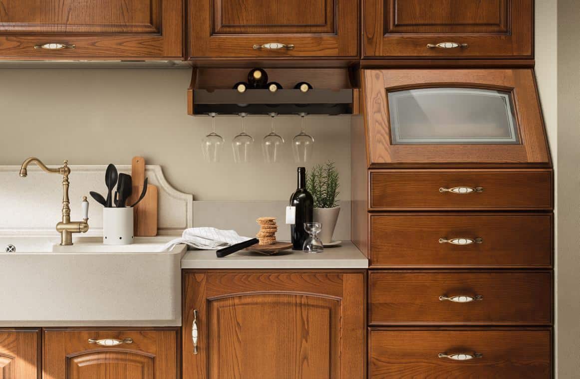 A kitchen with wooden cabinets, a farmhouse sink, a brass faucet, a wine bottle, wine glasses hanging upside down, a cutting board, kitchen utensils in a holder, and a small potted plant.