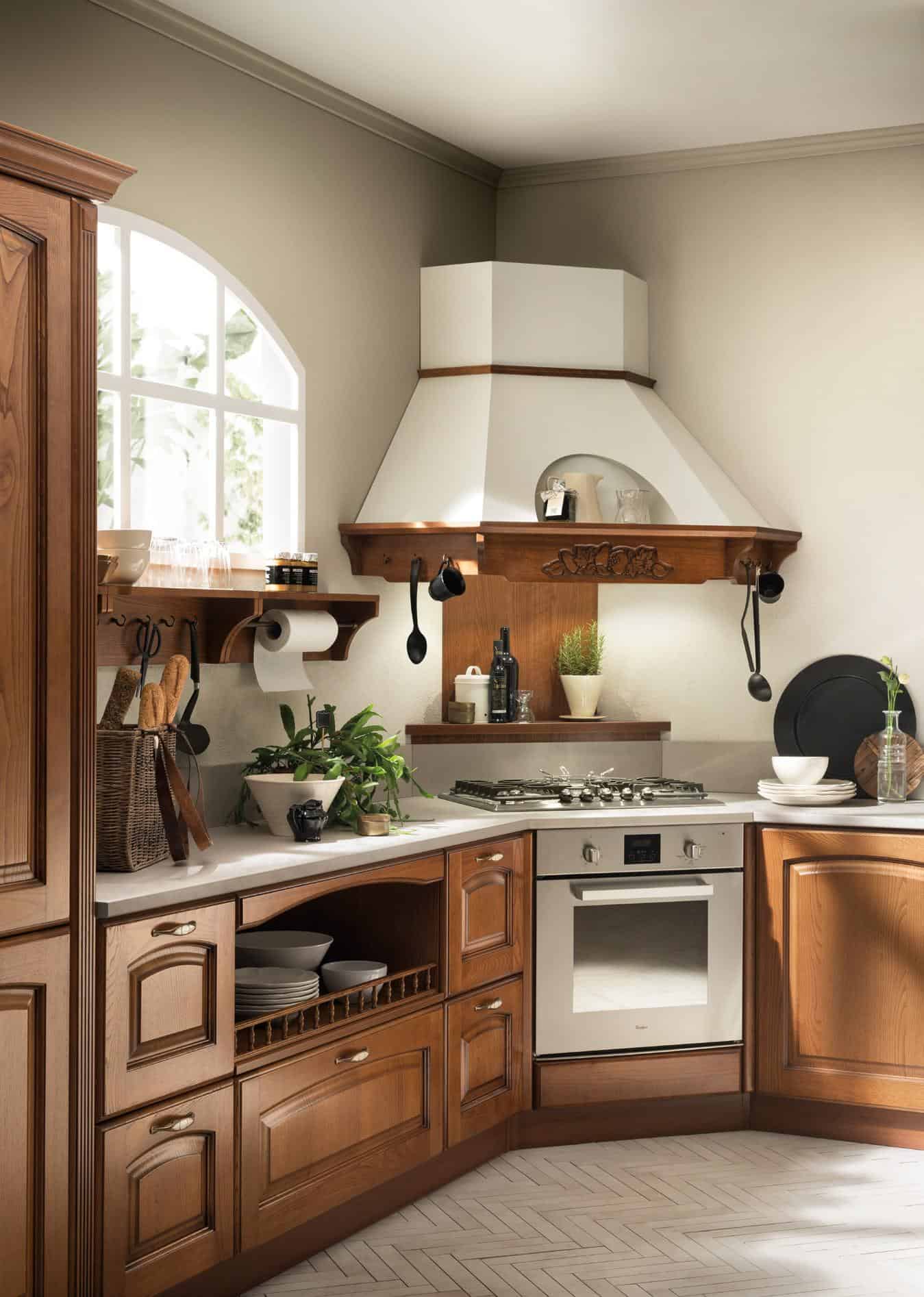 Traditional kitchen with wooden cabinets, a range hood, and built-in oven. There's a white countertop with plants, utensils, and bread in a basket. Light streams through an arched window, creating a warm atmosphere.