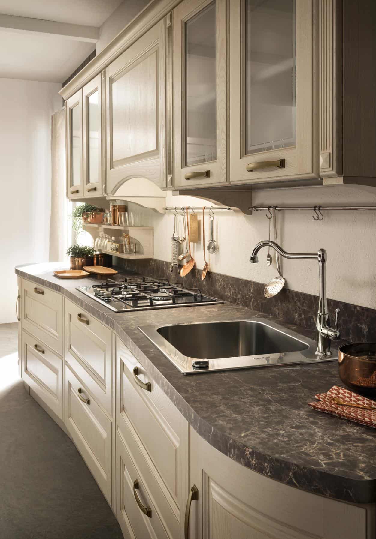 Modern kitchen with beige cabinetry, a marble countertop, stainless steel sink, and gas stove, featuring hanging utensils and copper accents.