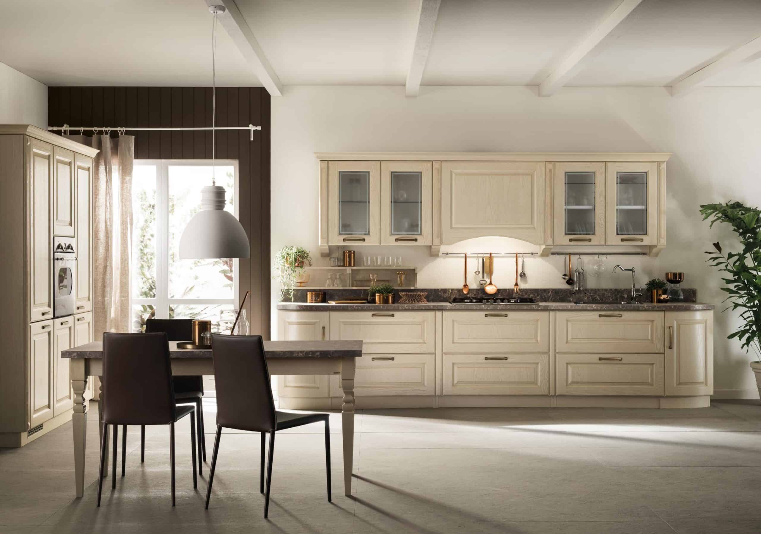Modern kitchen with light wooden cabinets, dark countertops, and a dining table with two chairs. A large window, overhead lighting, and a potted plant complete the setting.