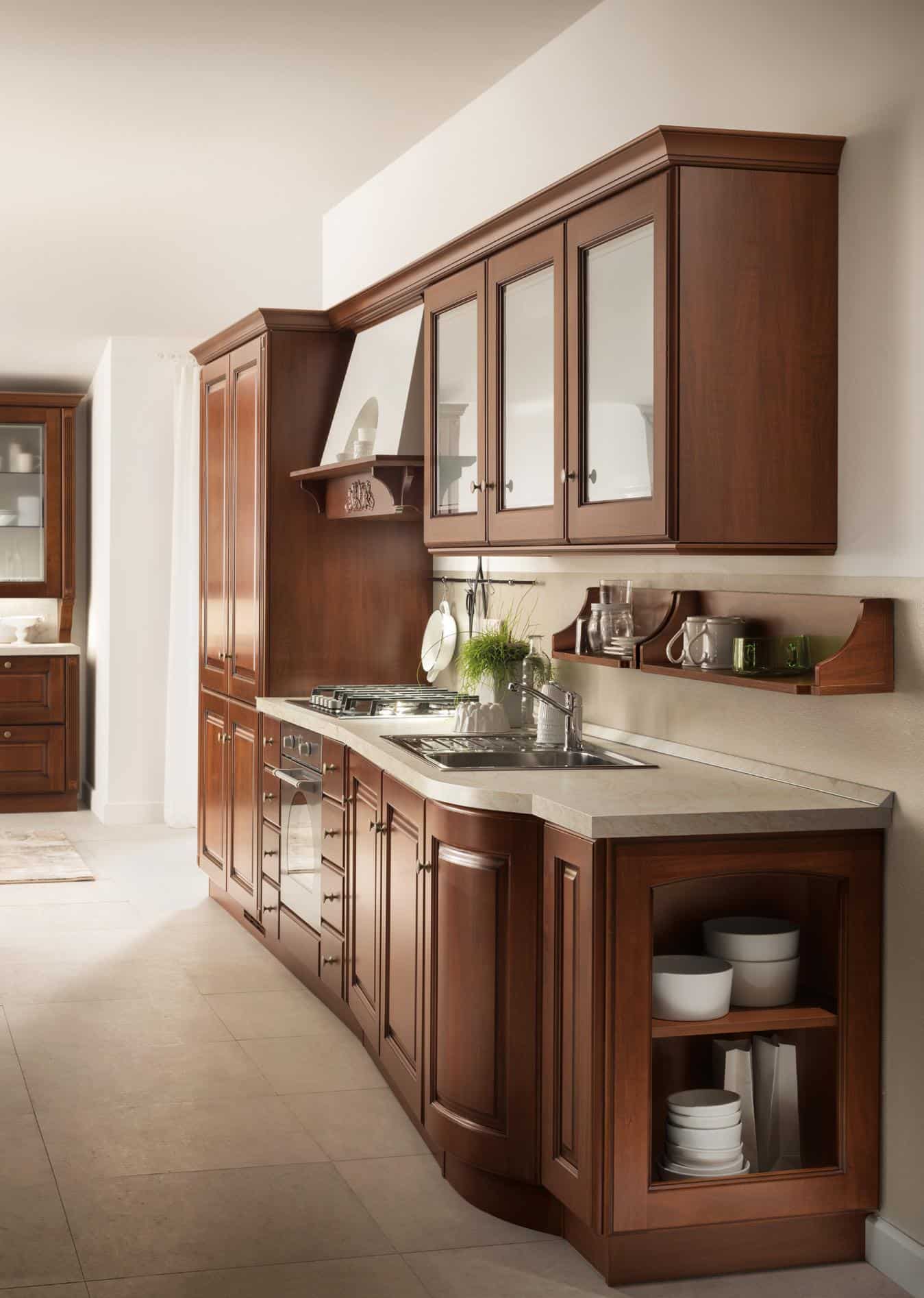 Traditional wooden kitchen cabinets with a marble countertop, featuring a stove, sink, and open shelves displaying dishes and plants.