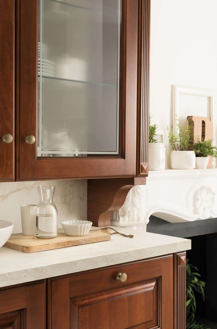 A kitchen setting with dark wooden cabinets featuring brass knobs, a glass-fronted upper cabinet, and a marble countertop. Decor includes a wooden tray with a glass carafe, a ceramic dish, and a white mug. A white decorative shelf with potted plants is in the background.