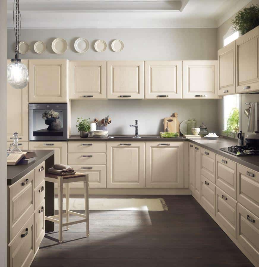 Modern kitchen with cream-colored cabinets, a dark countertop, and wooden flooring. Pendant light hangs over an island with a stool. Decor includes plants and plates on the wall.