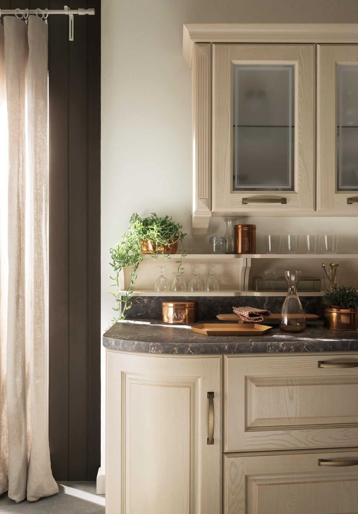 Light wood kitchen cabinets with glass-paneled doors, dark countertop, potted plants, copper containers, and hanging glasses under soft natural light.
