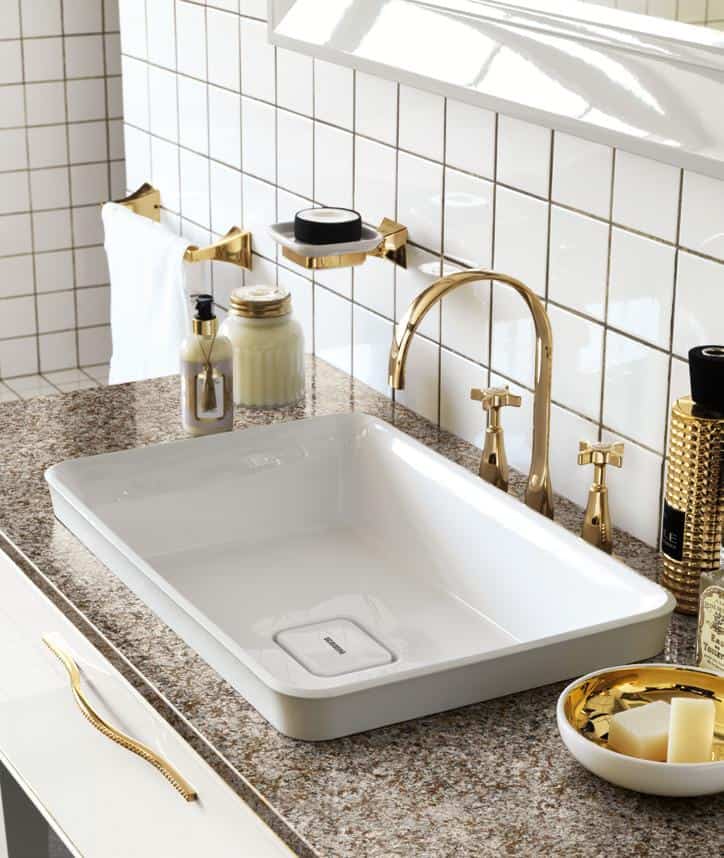 A modern bathroom sink with a gold faucet, surrounded by toiletries on a granite countertop. The wall features white square tiles.