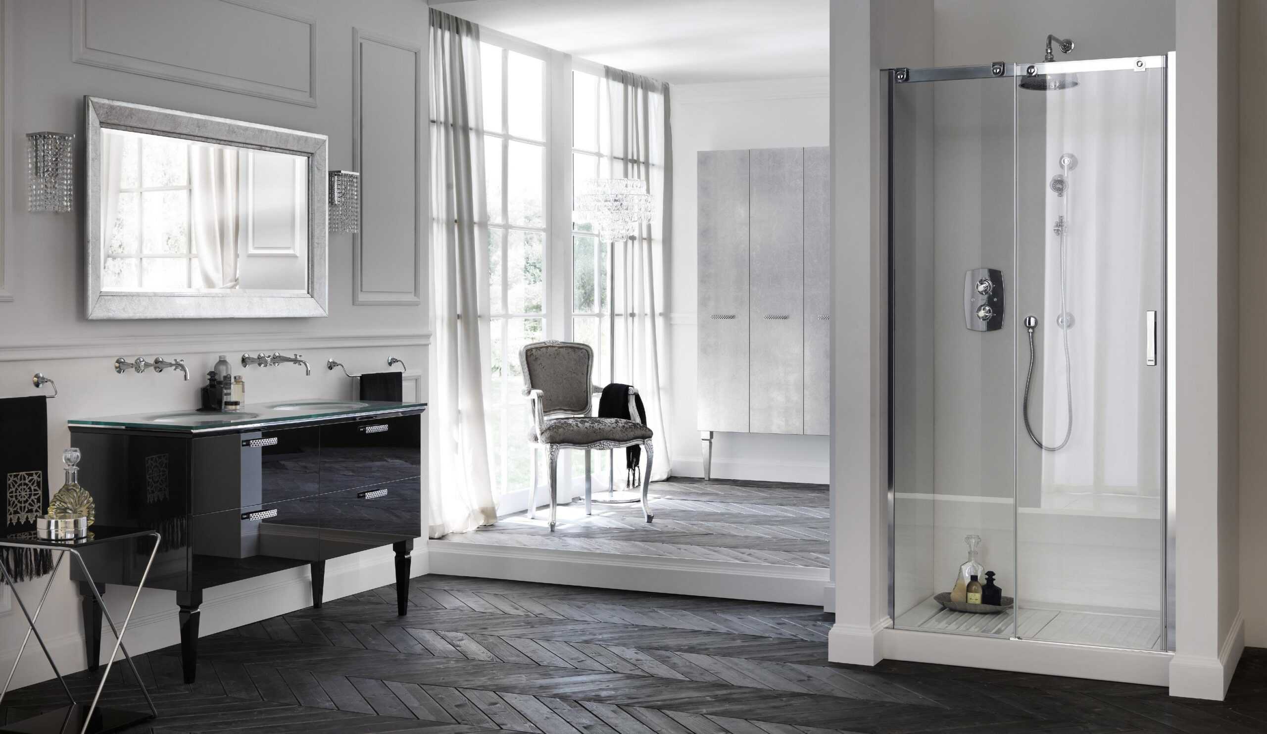 Elegant bathroom with a glass-enclosed shower, black vanity, large mirror, cushioned chair, and pendant light, featuring chevron-patterned wooden flooring and floor-to-ceiling windows with sheer curtains.
