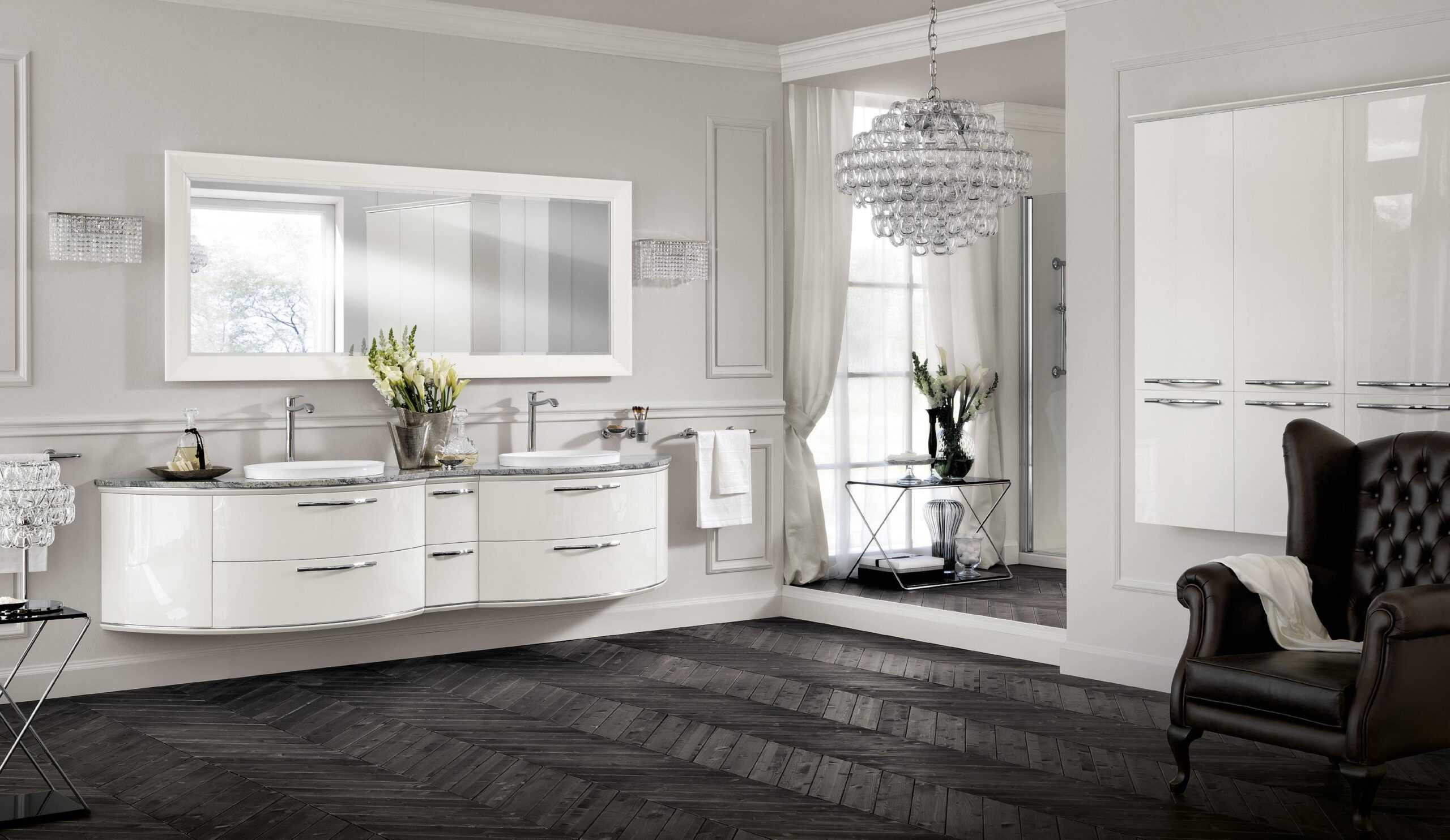 Modern bathroom with double sinks, white cabinets, a wall mirror, chandelier, dark herringbone floor, and a leather armchair.