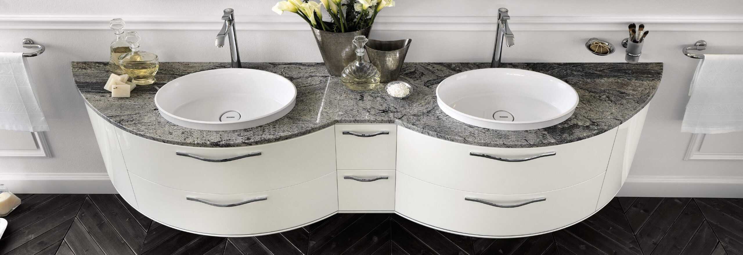 Double sink bathroom vanity with a curved design, featuring a granite countertop and white basins, complemented by chrome faucets and elegant handles.