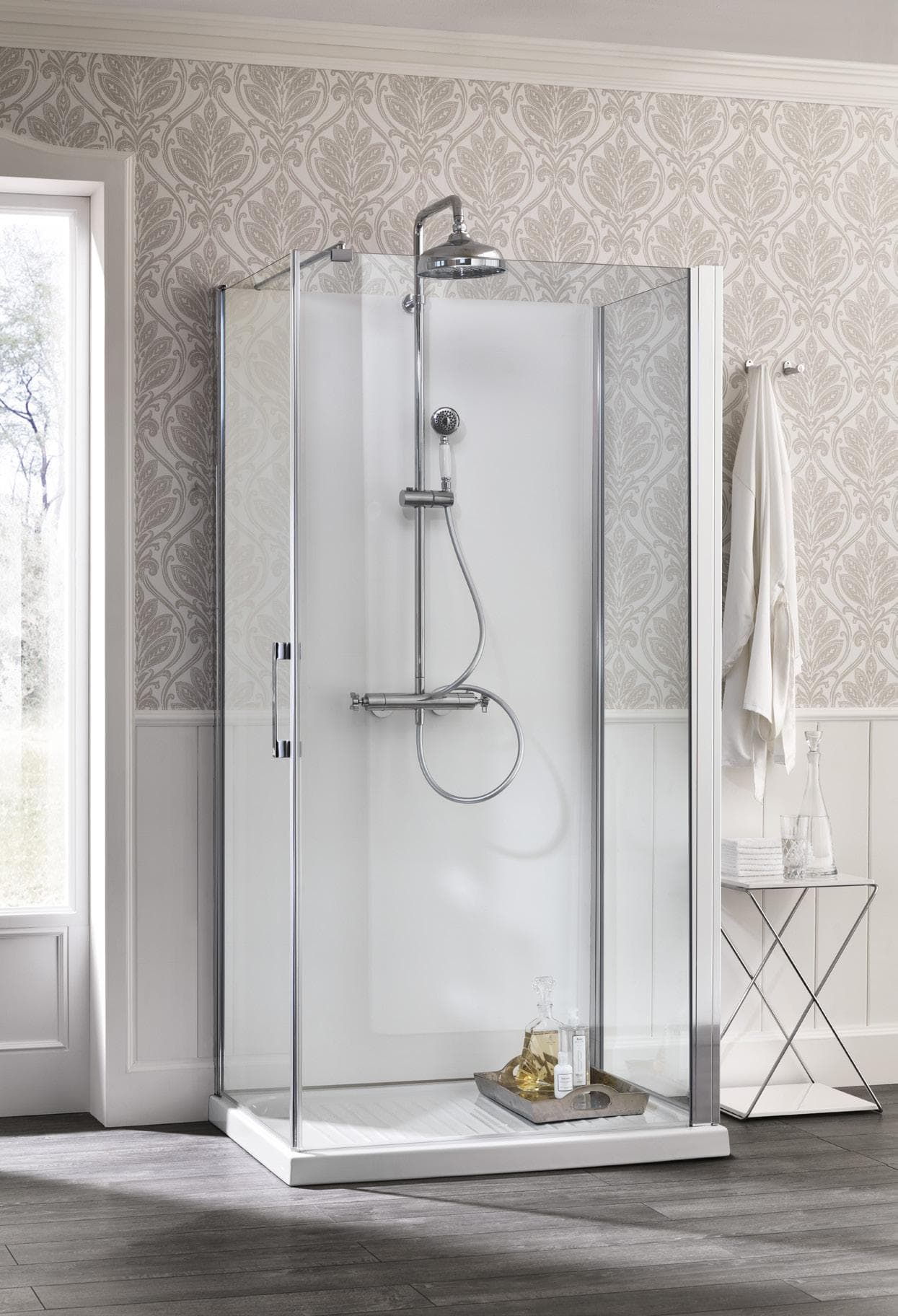 Glass shower enclosure with silver fixtures in a bathroom with patterned wallpaper and wooden flooring.