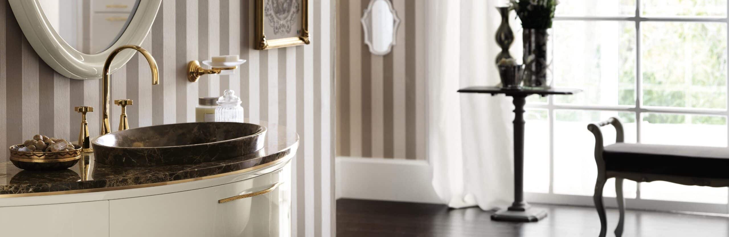 Elegant bathroom with a marble sink and brass fixtures against striped wallpaper, featuring a large mirror and a side table near a window with sheer curtains.