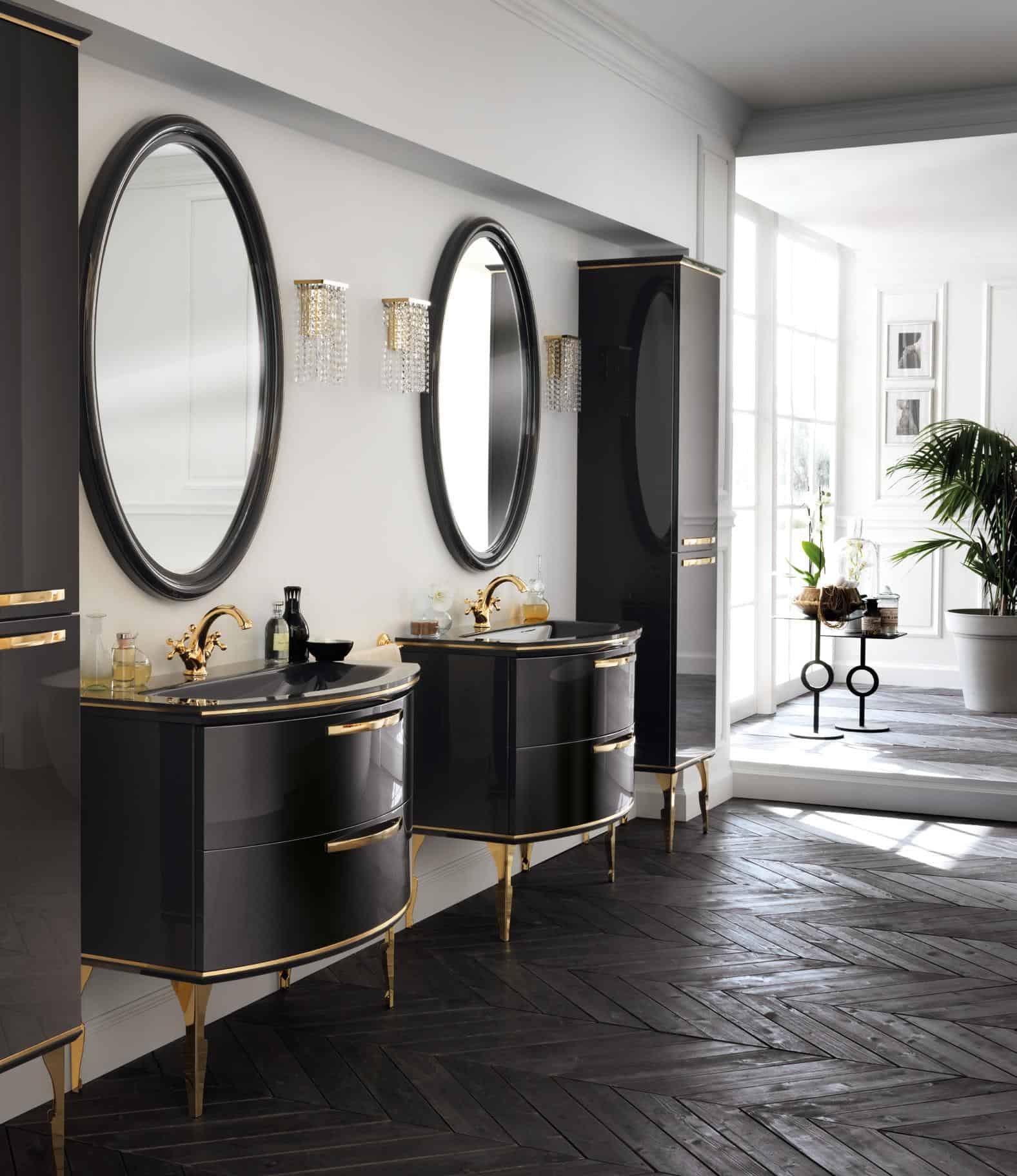 Luxurious bathroom with two black vanities featuring gold accents, oval mirrors above each vanity, crystal wall sconces, dark herringbone wood flooring, and large windows with natural light.
