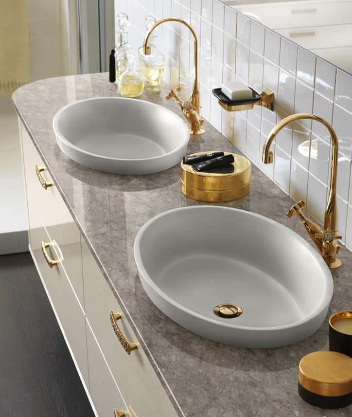Modern bathroom countertop with two oval sinks, gold faucets, and decorative gold-accented items.