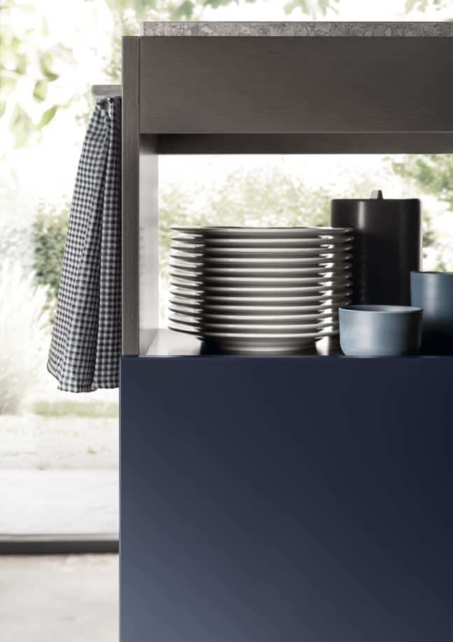 A stack of white plates, a dark pitcher, and a blue cup on a shelf with a checkered cloth hanging on the side.