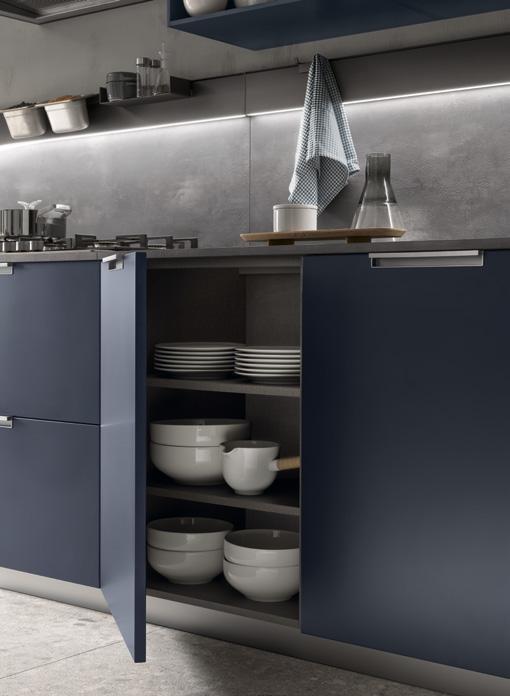 Modern kitchen with dark blue cabinets, open door revealing stacked white dishes and bowls, stainless steel countertop with a kettle, and a checkered cloth hanging above.